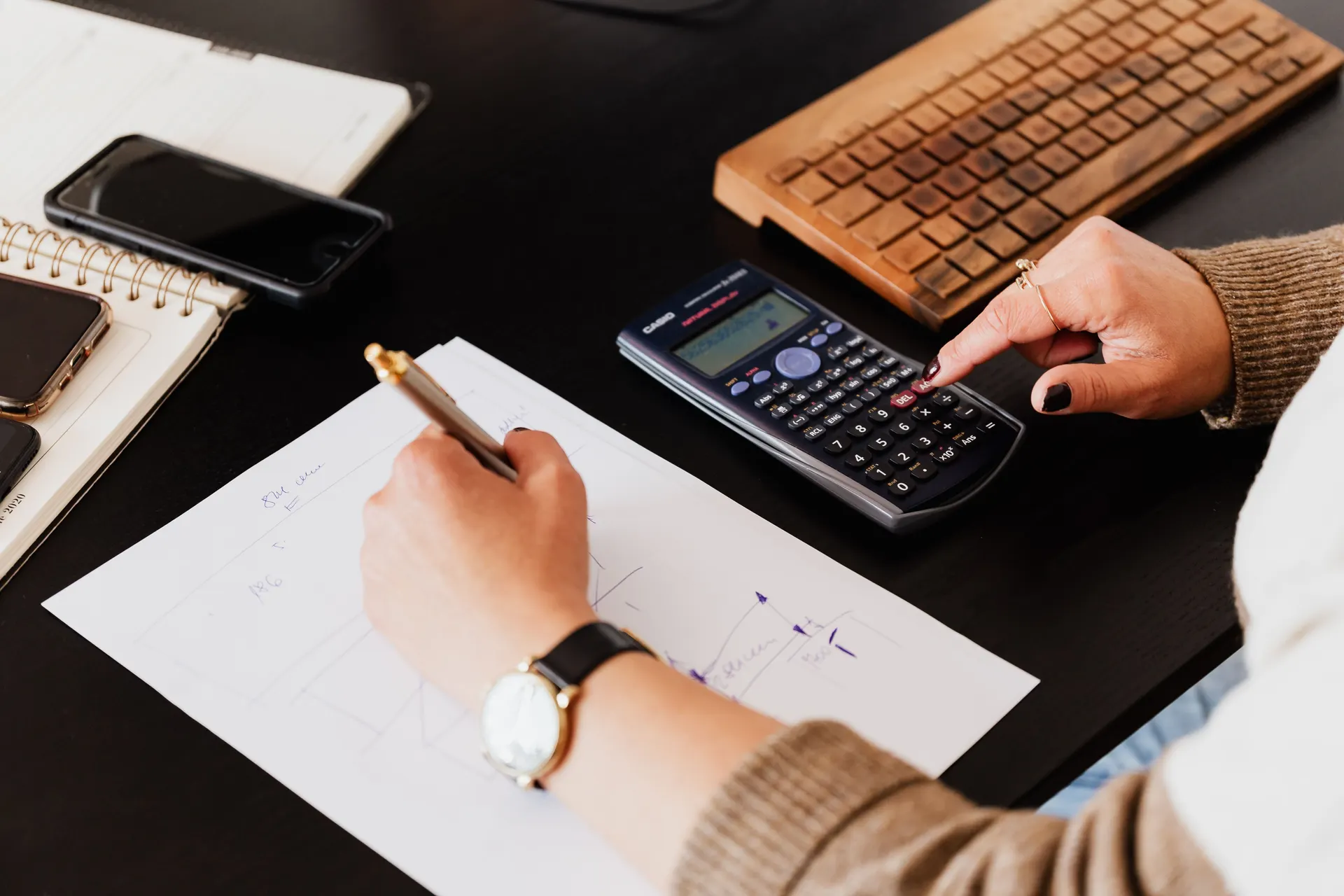 Hands at a desk working a calculator and writing figures on paper, a phone and notebook nearby