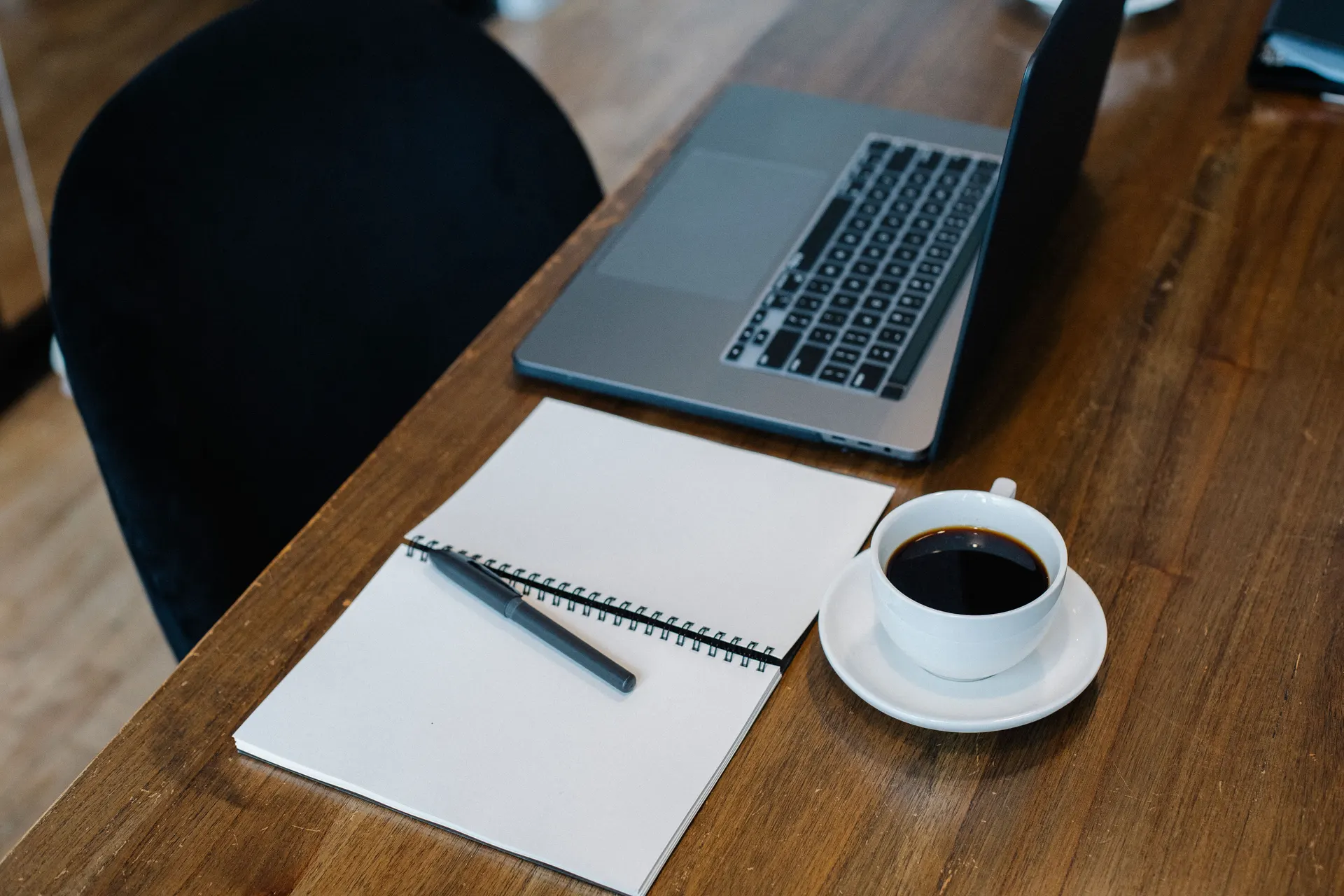 A desk in window light with an open laptop, a notebook and a cup of coffee